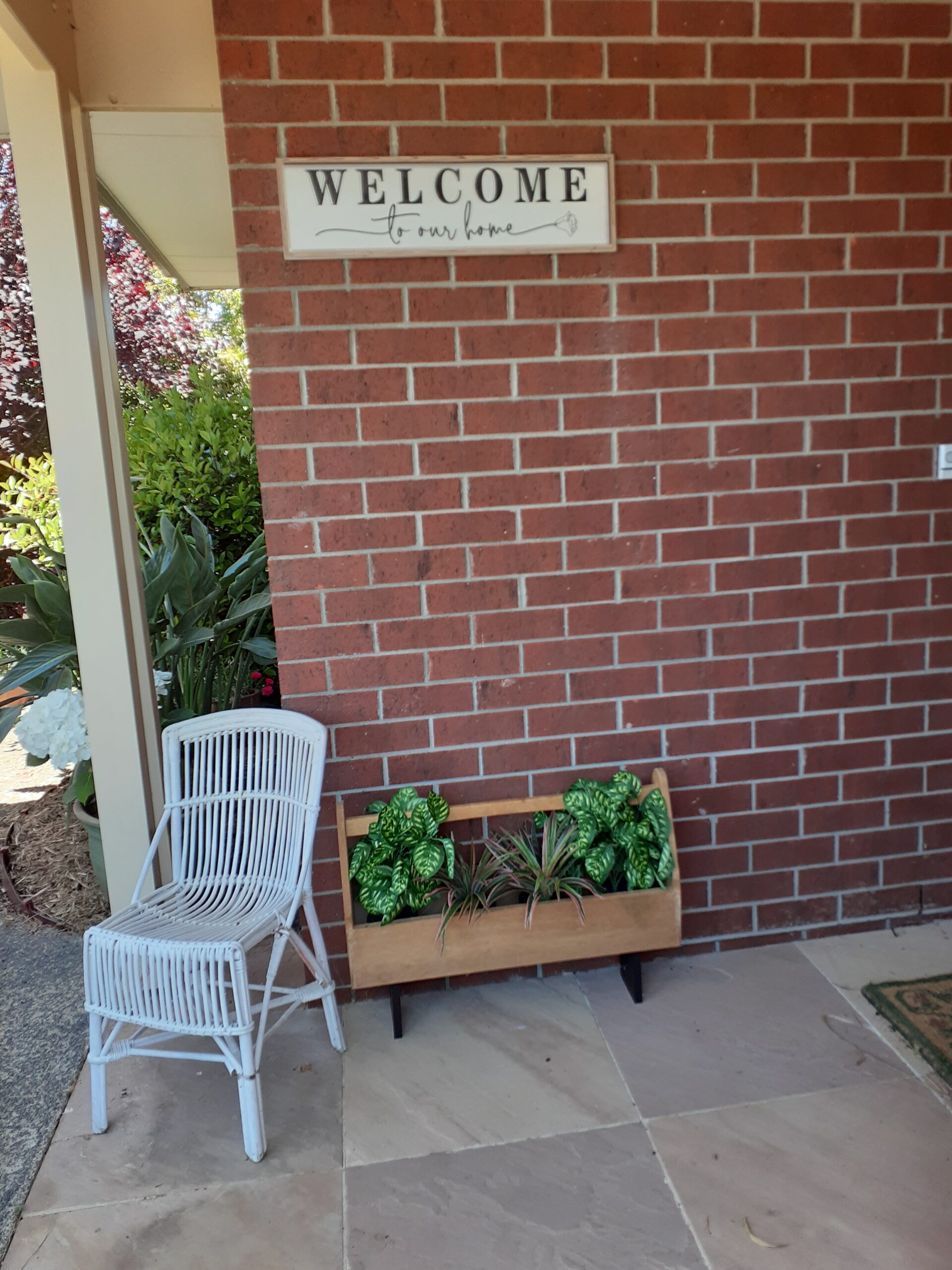 photo of a sign saying welcome to my home on the brick wall of my front porch, above a pot plant and cane garden chair